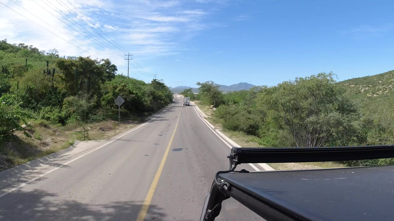 Vehicle-mounted view along a straight Los Cabos road, with a yellow center line, scrub and trees on both shoulders, utility wires crossing the upper left, distant hills, and no visible people.
