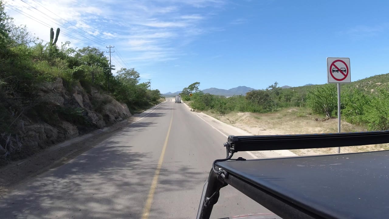 The Los Cabos roadway continues straight ahead, with the sign still visible at right, rocky vegetation at left, distant hills, and no visible people.