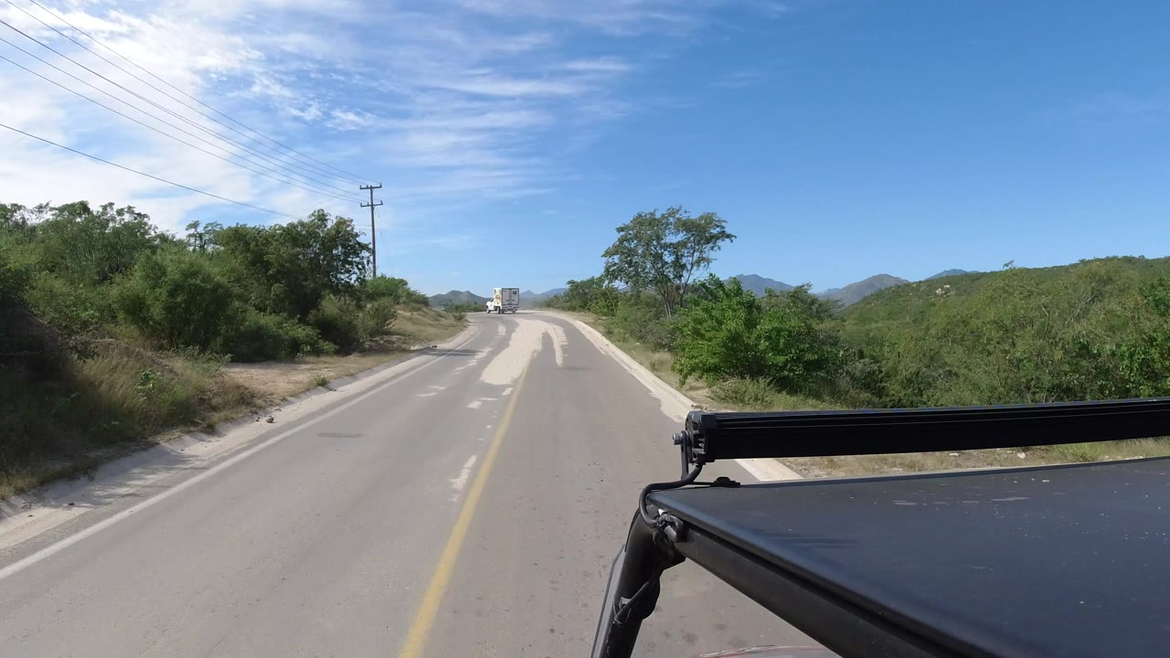 Forward view from a black vehicle on a Los Cabos road, with scrubby green hills, utility lines, a bright blue sky, and no visible people.