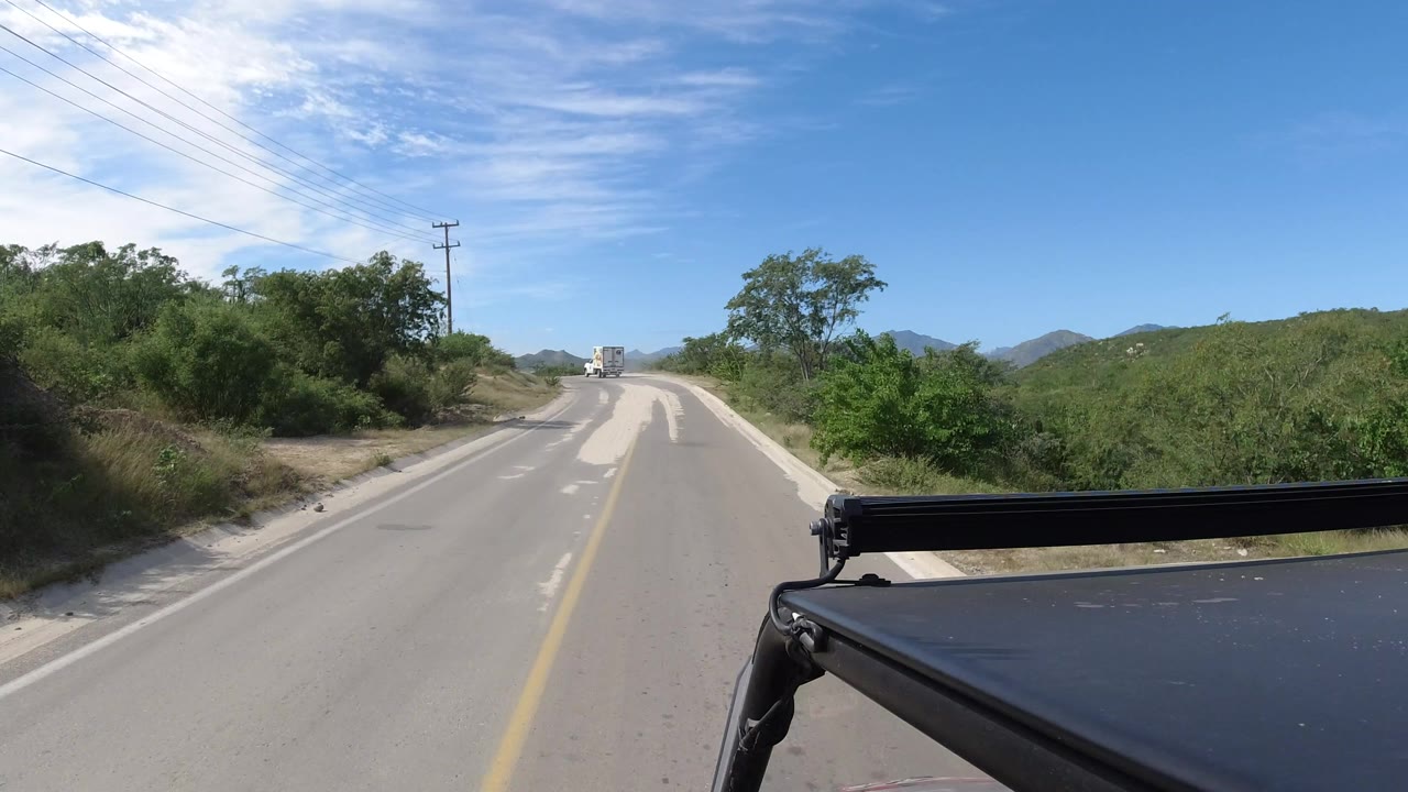 A black vehicle foreground frames a sunlit Los Cabos road continuing between green roadside brush and distant hills, with no visible people.