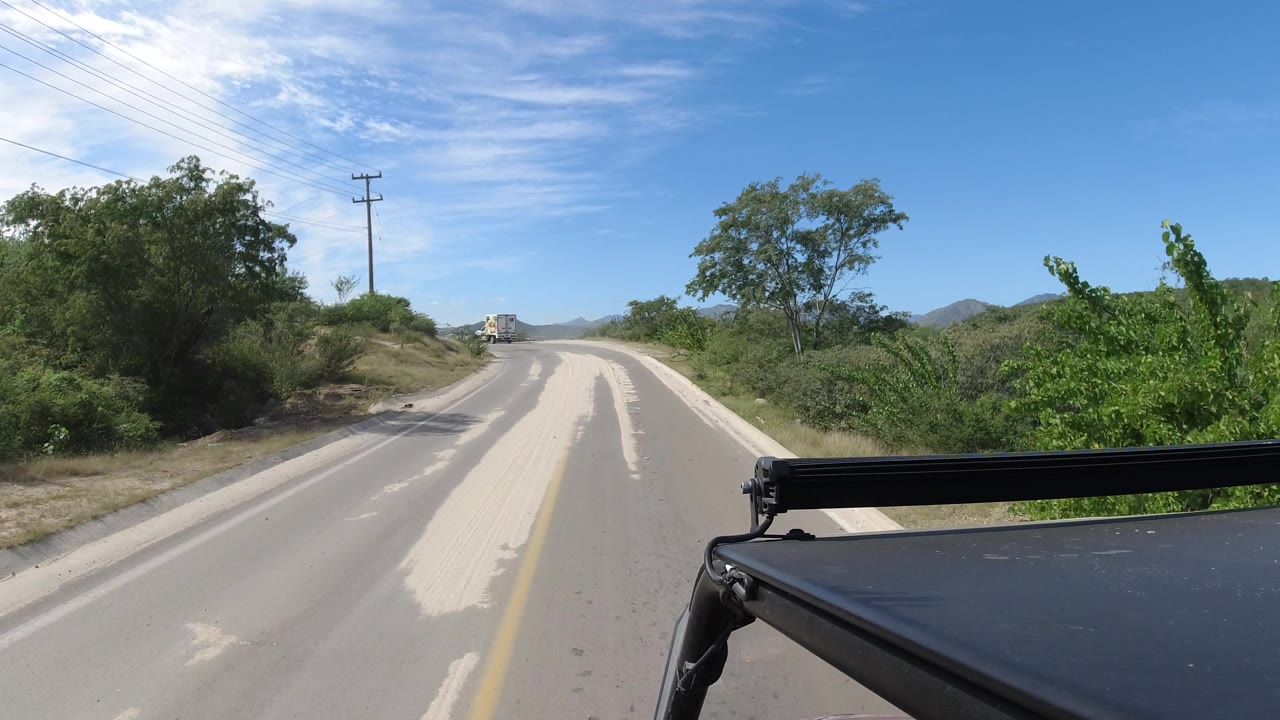 Curving road with pale sand across the pavement and shrubs on both sides.
