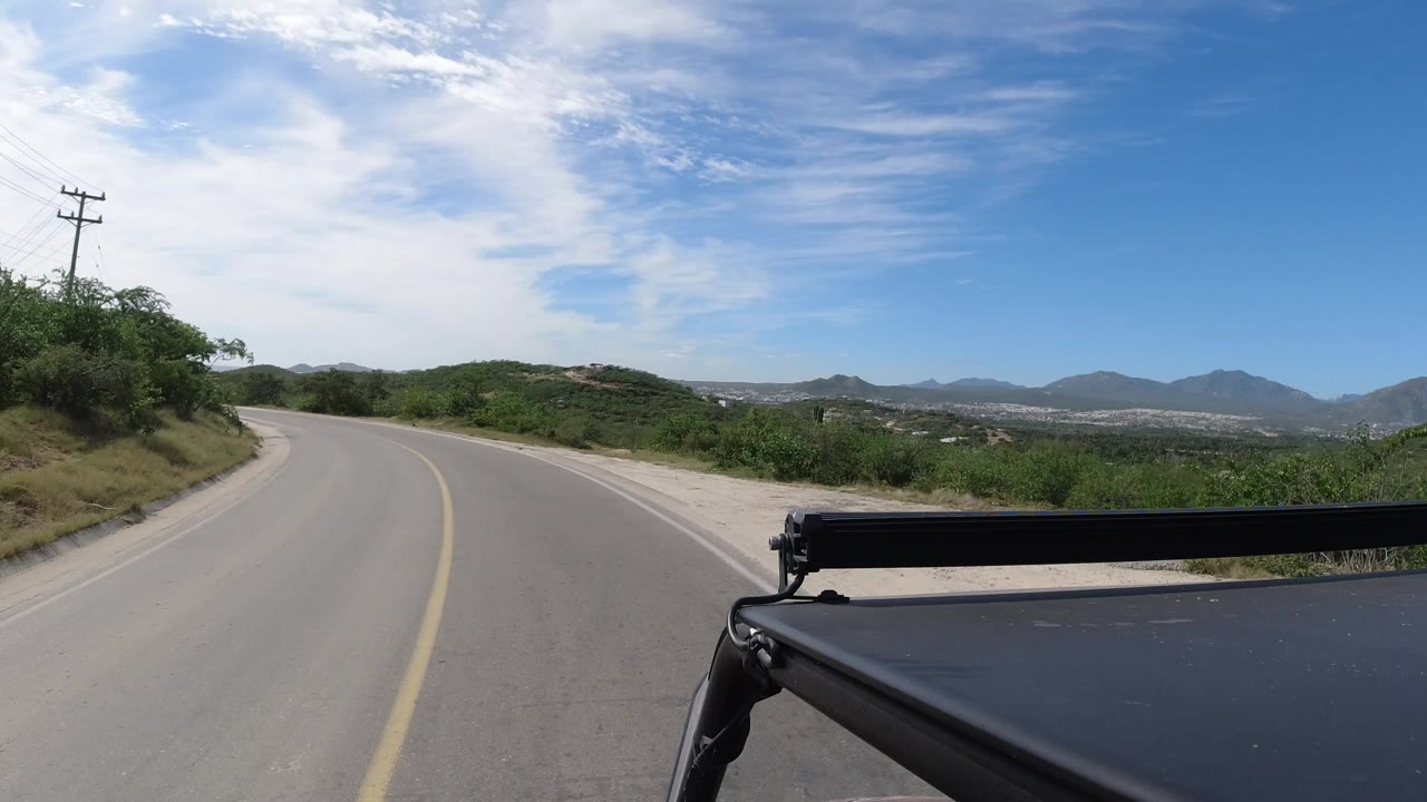 Curving paved road with green brush and distant hills under a blue sky.