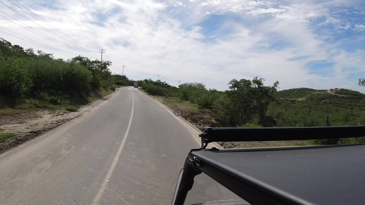 Forward view down a quiet paved road with shrubs and a vehicle roof in foreground.