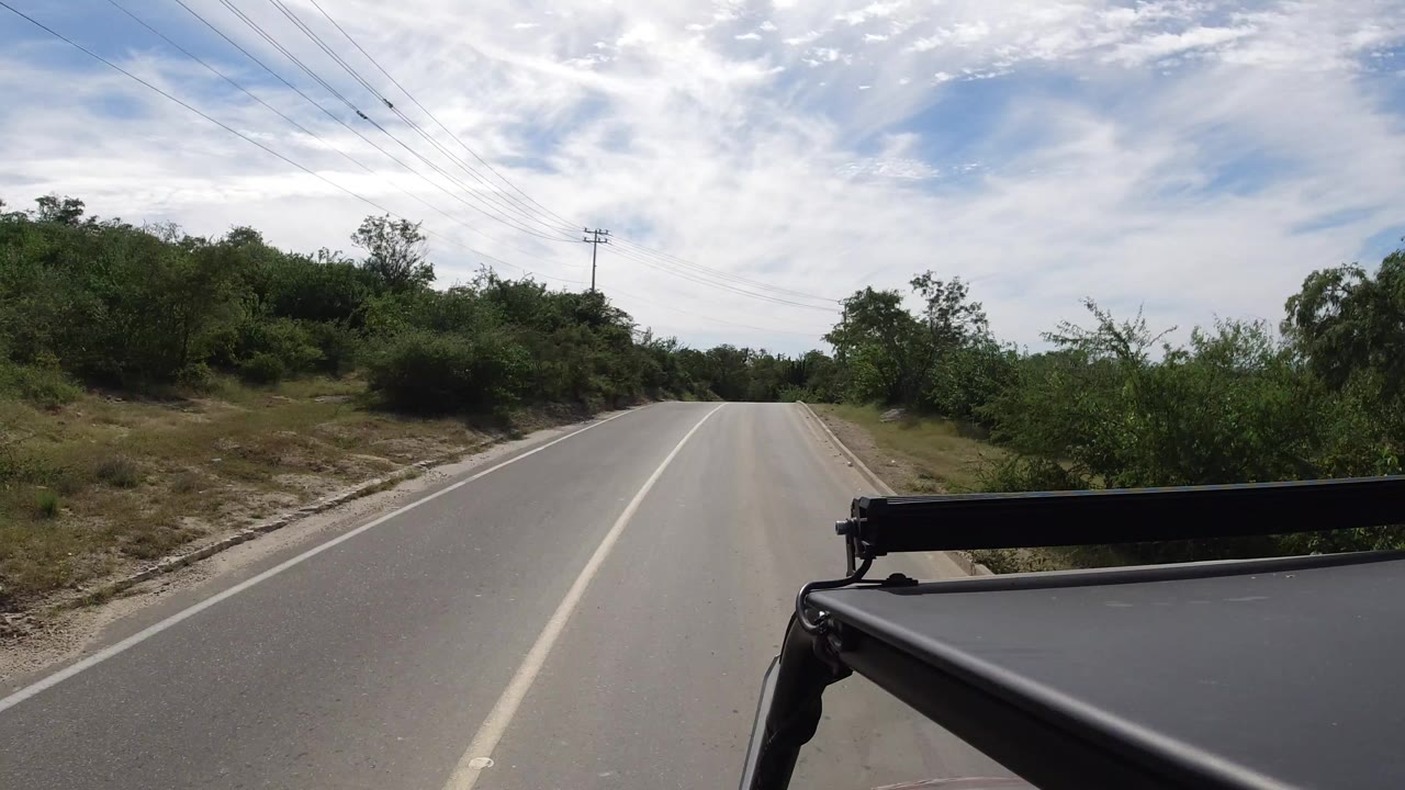 Forward view from a vehicle on an empty paved road in Los Cabos, Mexico, with green bushes on both sides, overhead wires, cloudy blue sky, and no visible people.