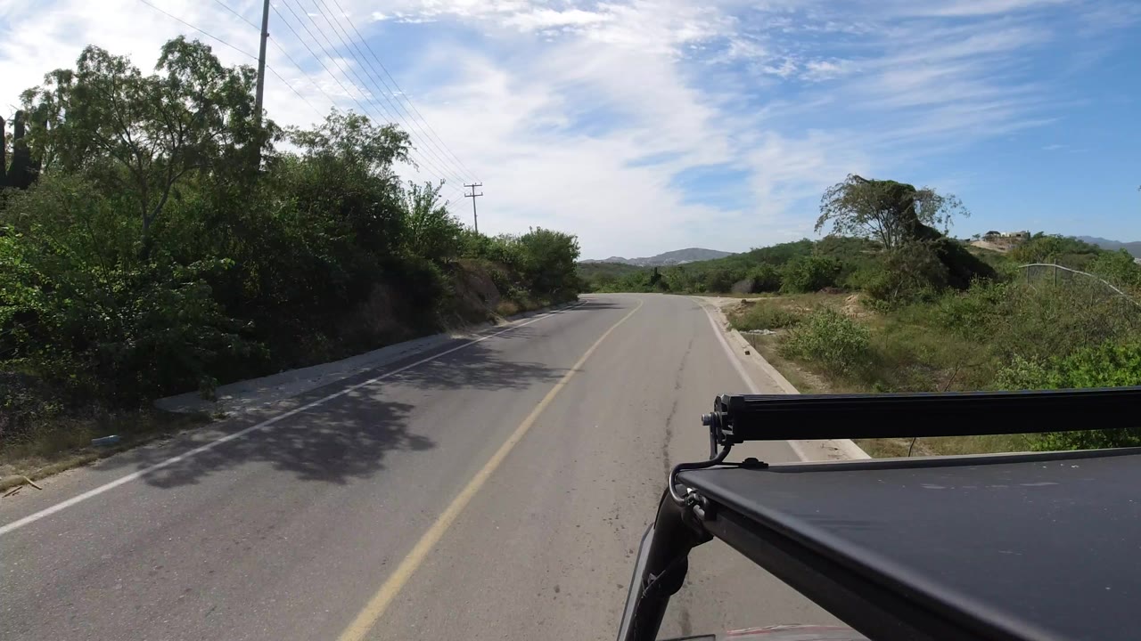 The road continues through Los Cabos, Mexico with a centered yellow line, thick shrubs along both shoulders, distant hills, a bright clouded sky, and no visible people.