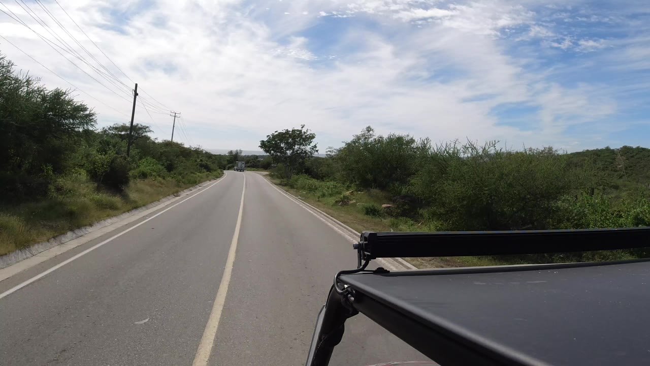 A Los Cabos, Mexico paved road runs forward between green brush and utility poles under a bright cloudy sky, with a dark vehicle edge in the lower right and no visible people.
