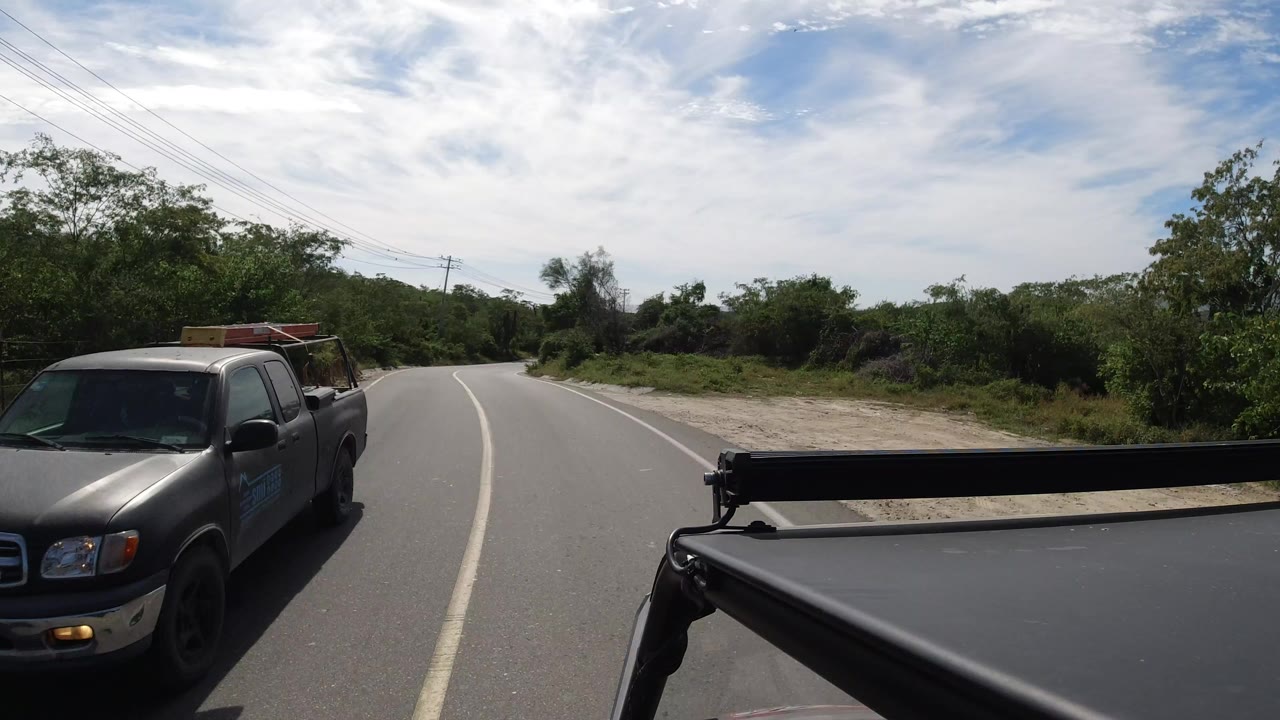 The Los Cabos road view continues past a pickup truck on the left, with the camera vehicle's black support bar in front, scrubby green vegetation, utility wires, and no visible people.