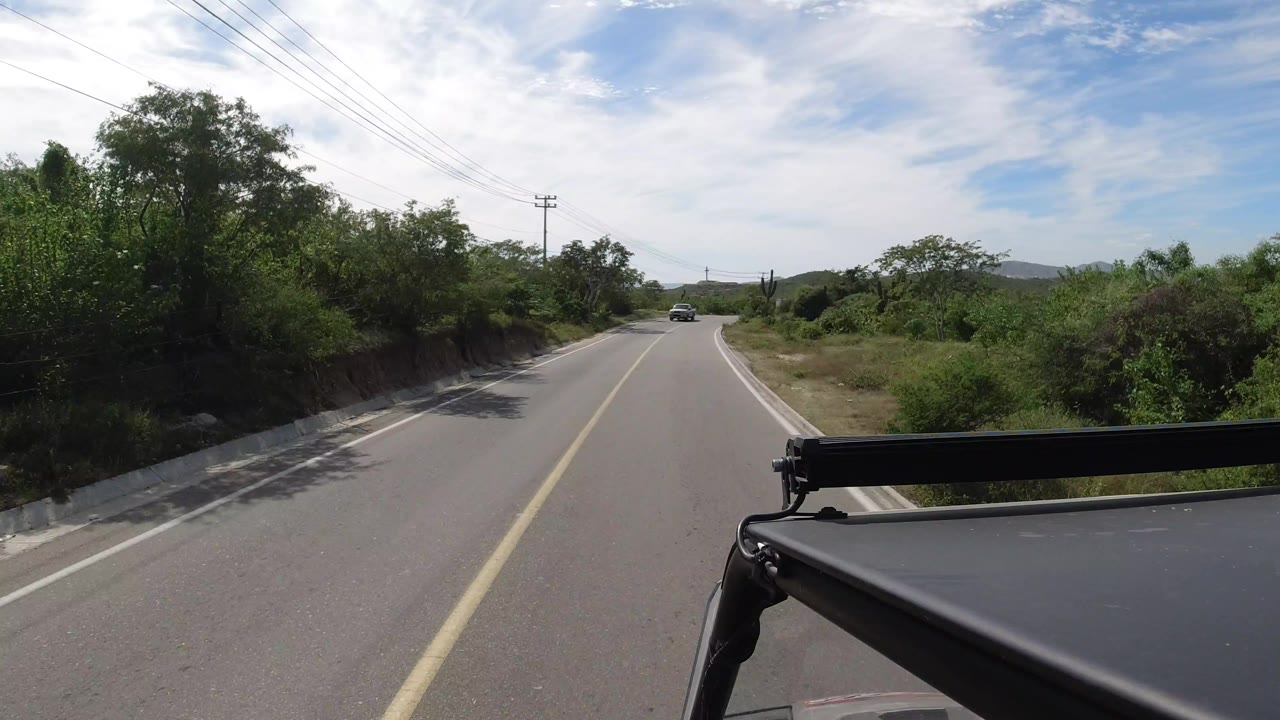 Los Cabos road scene with the center line leading toward a bend, dense green roadside vegetation, overhead utility wires, low hills, foreground vehicle hardware, and no visible people.
