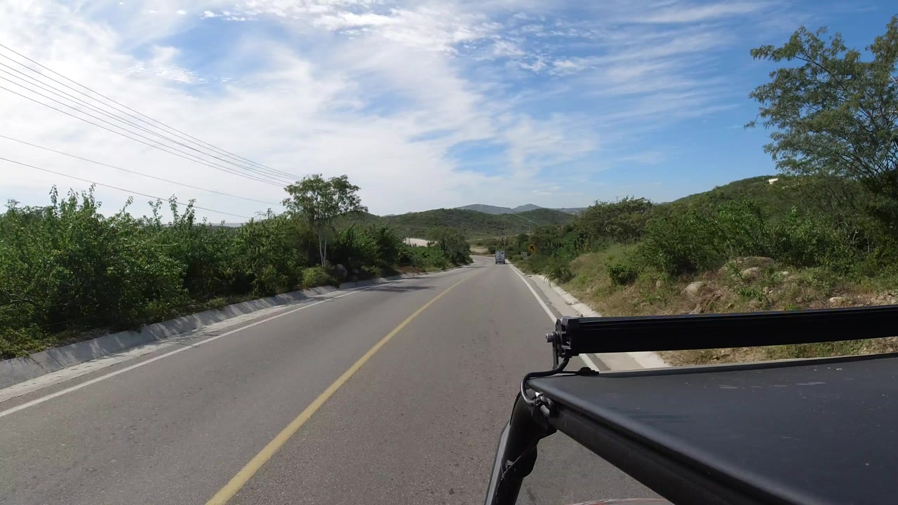 Opening view down a two-lane road in Los Cabos, Mexico, with roadside shrubbery, low hills, utility lines, and part of a black vehicle rack visible, with no visible people.