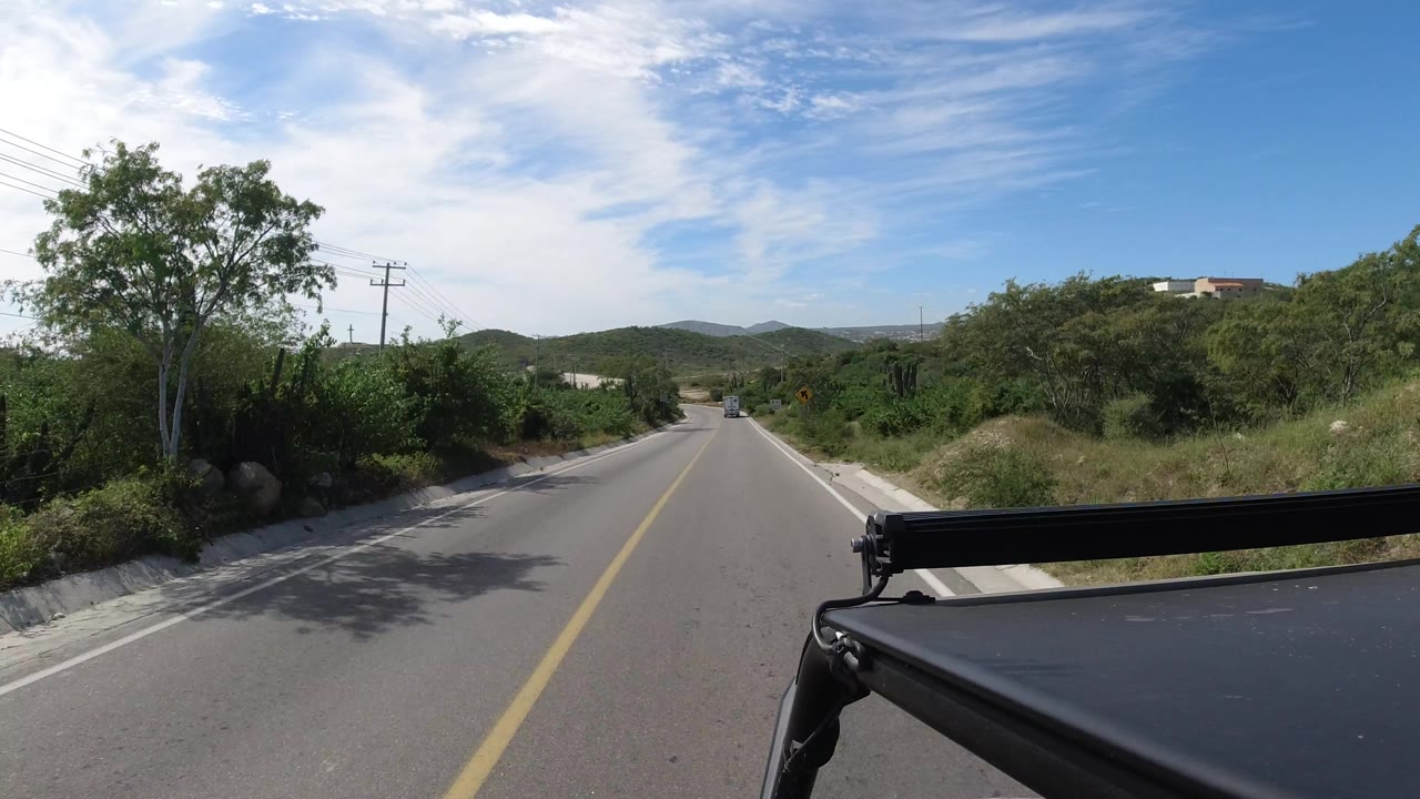 The road in Los Cabos, Mexico, bends slightly past a leafy tree, roadside bushes, low hills, and utility poles, with part of the vehicle rack in frame and no visible people.