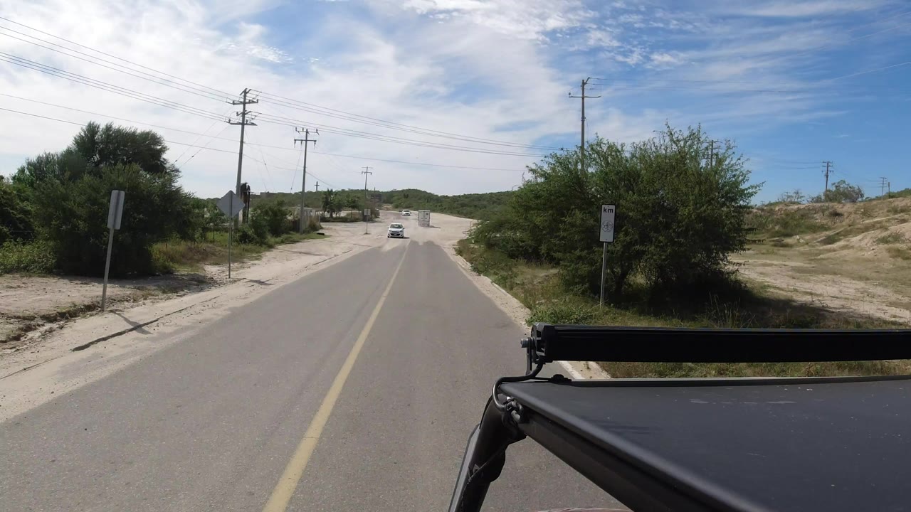Opening view of a Los Cabos paved road with low green shrubs, utility poles, overhead wires, distant hills, a black vehicle edge at the bottom, and no visible people.