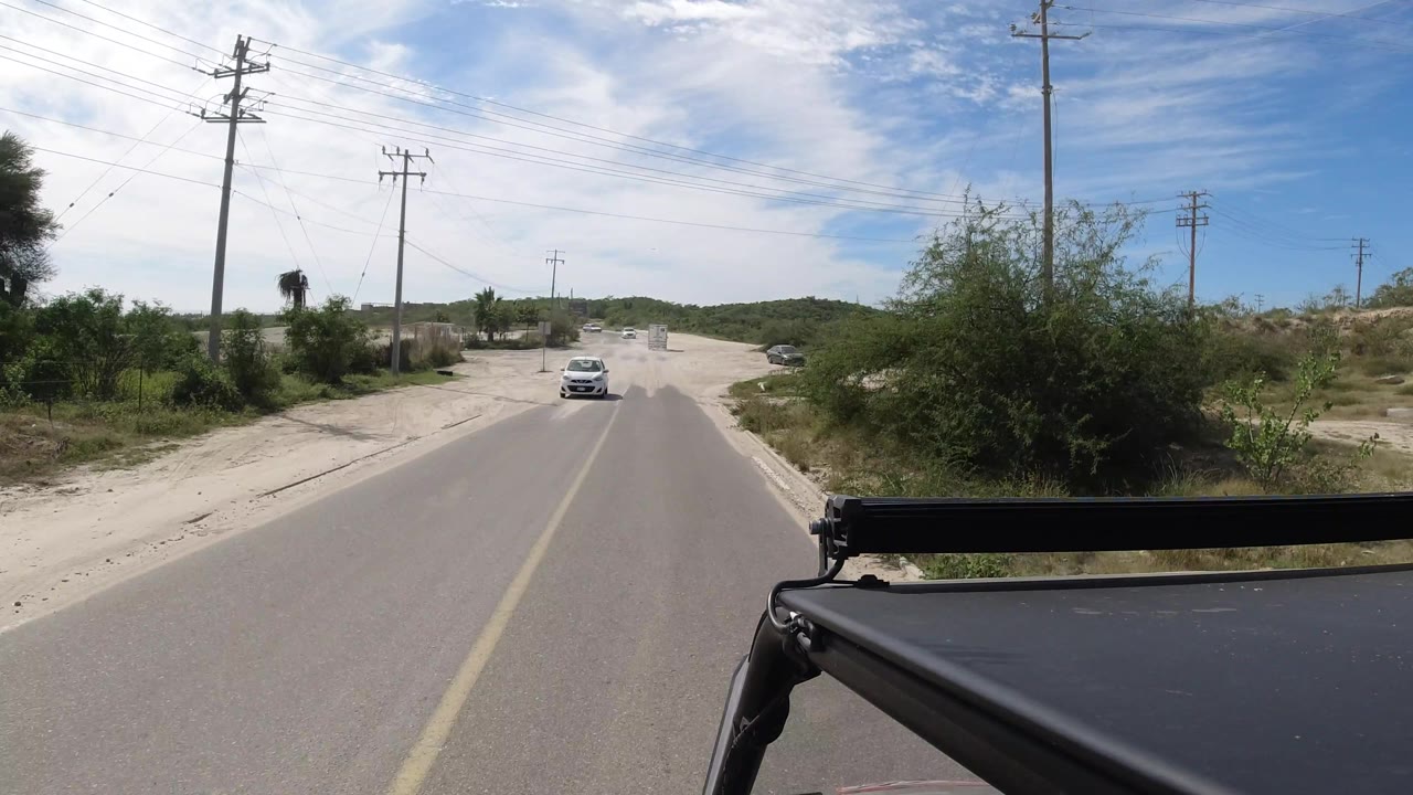 White car approaching on a road bordered by sand and shrubs.