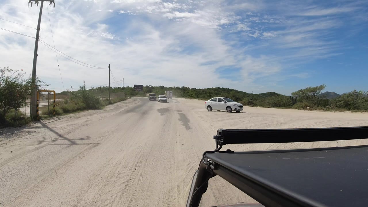 Sandy road with cars, utility poles, brush, and blue sky.