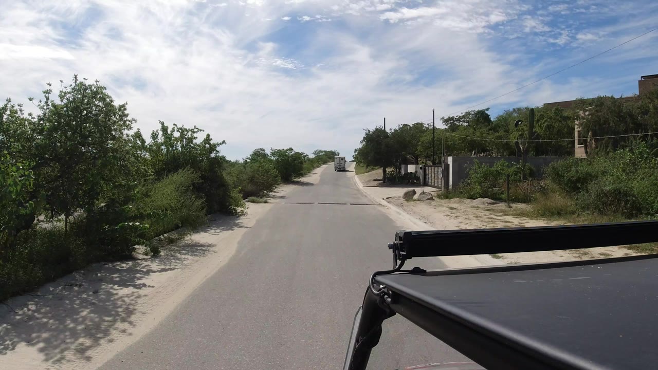The road remains centered in Los Cabos with a distant truck ahead, dense shrubs at the edges, a black rack across the lower frame, and no visible people.