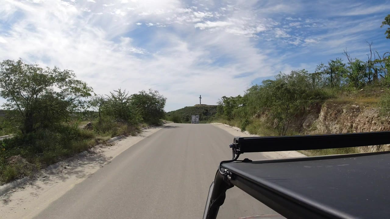 Forward view along a small Los Cabos road bordered by dense bushes, with a distant hilltop cross-like structure, vehicle roof edge in the foreground, and no visible people.