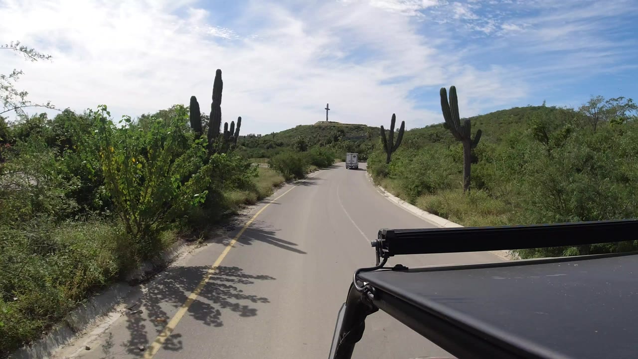 Forward road view in Los Cabos, Mexico, with tall cactus on both sides, a small truck in the distance, a hilltop cross, blue sky, and no visible people.
