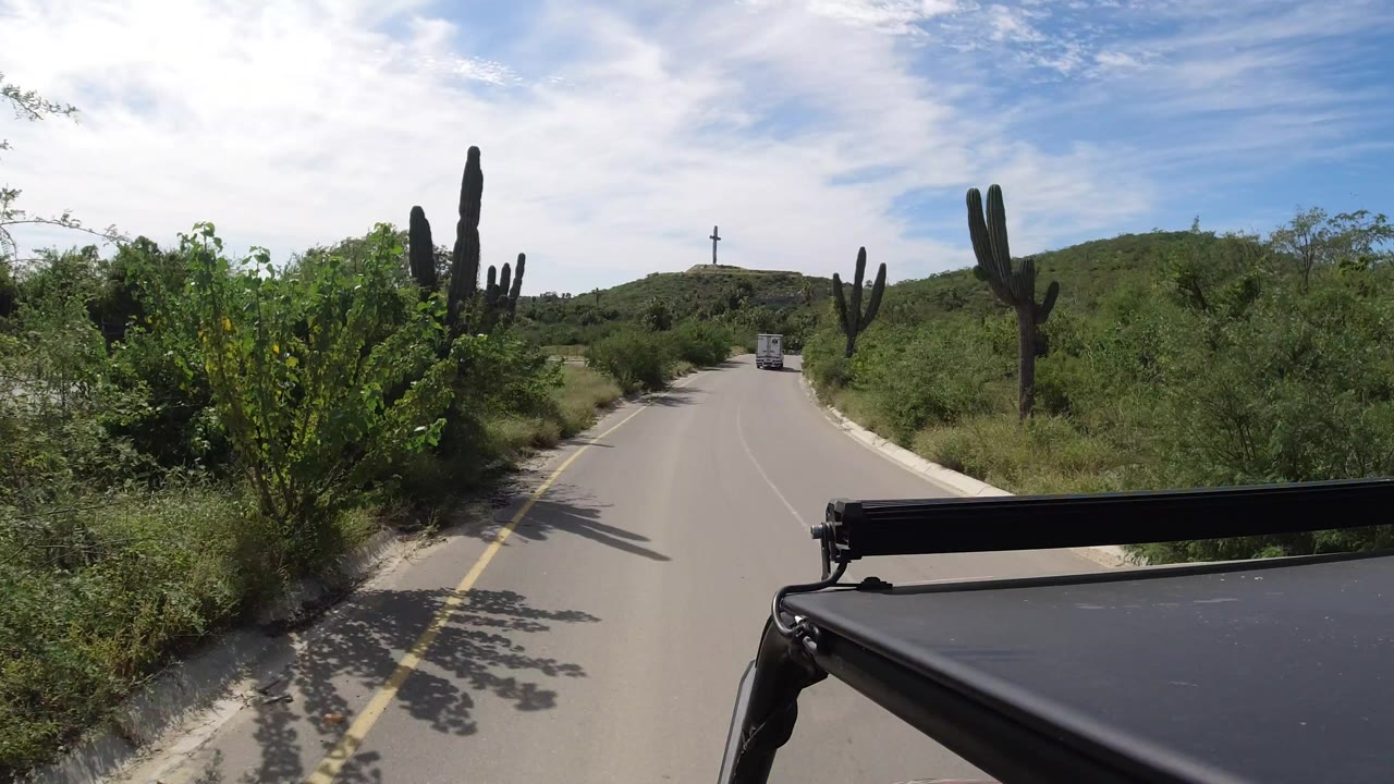 Forward-facing road frame in Los Cabos, Mexico, showing the vehicle continuing past cactus, dense shrubs, a distant truck, low hills, and no visible people.