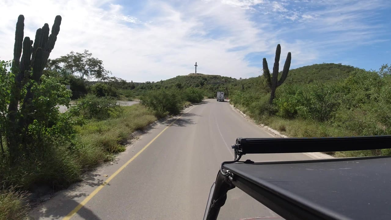 Road passing a tall cactus with hills and a small truck ahead.