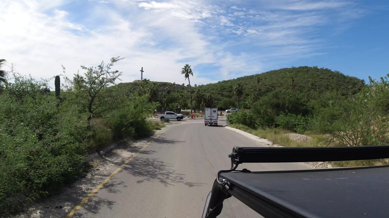 A vehicle-mounted Los Cabos roadway frame shows the white truck farther ahead, a roadside parking area, palms, scrub vegetation, low hills, and no visible people.