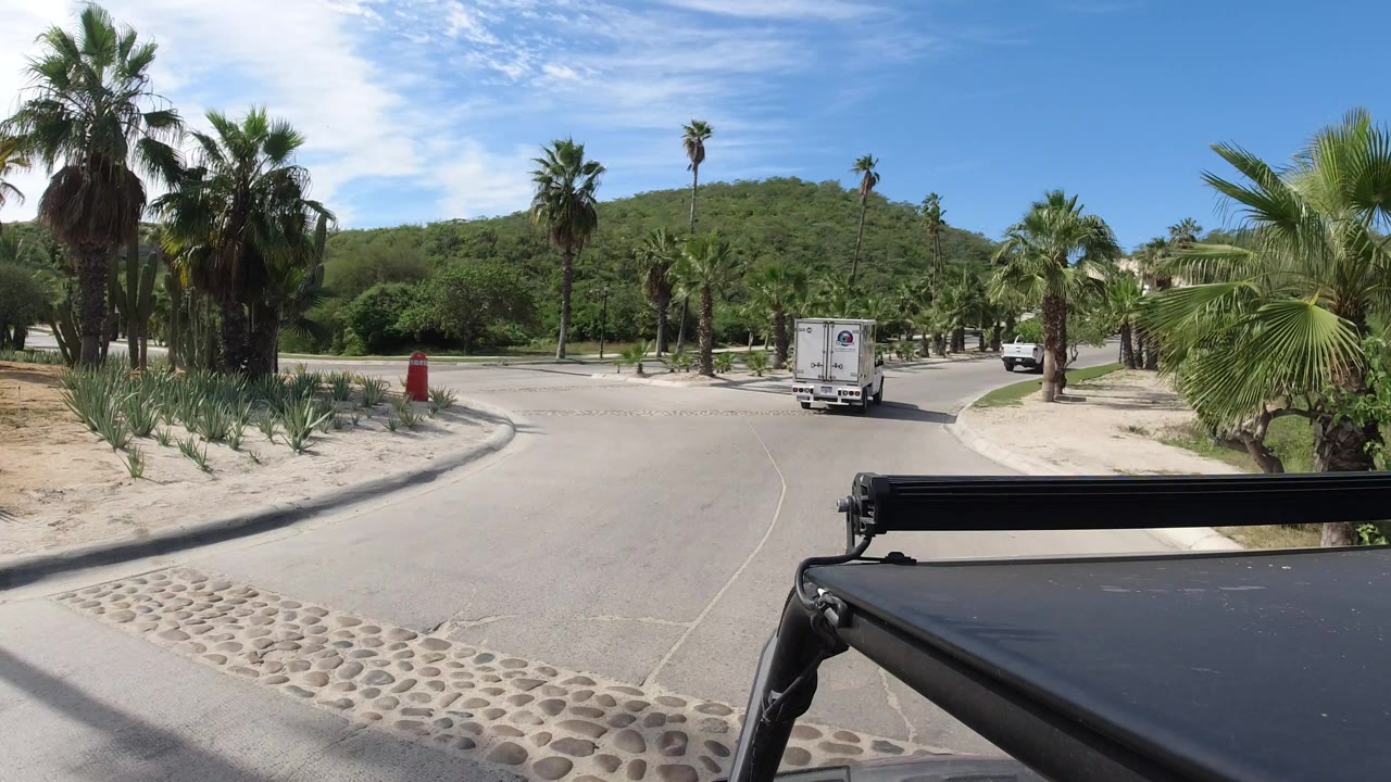 One second in, the white delivery truck continues away along the Los Cabos road as palms, low hillside greenery, and a bright sky fill the background, with no visible people.