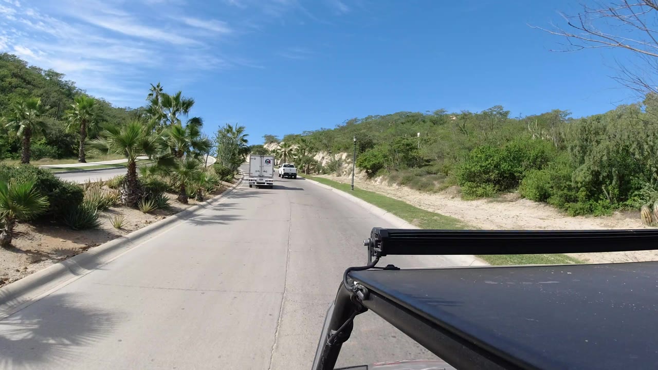 The next frame keeps the white vehicle centered on the paved road, with palms, dry shrubs, sandy banks, and no visible people in Los Cabos.