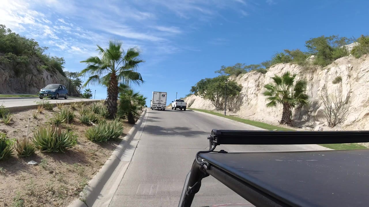 A Los Cabos road scene shows cars ahead, a palm and desert plants beside the curb, pale rock hills, blue sky, and a dark vehicle rail in the foreground, with no visible people.