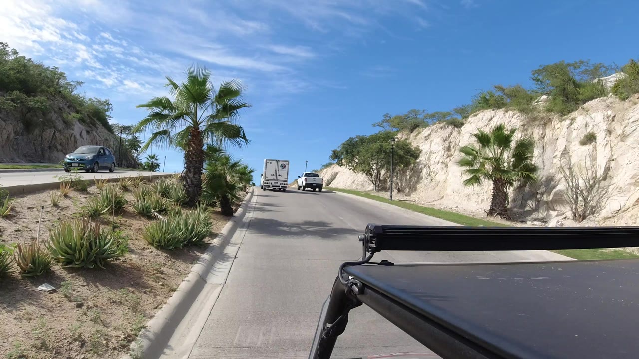 A Los Cabos roadway continues uphill between planted palms, sandy roadside beds, pale cut hills, and vehicles ahead, with a dark vehicle roof or rail at the bottom and no visible people.