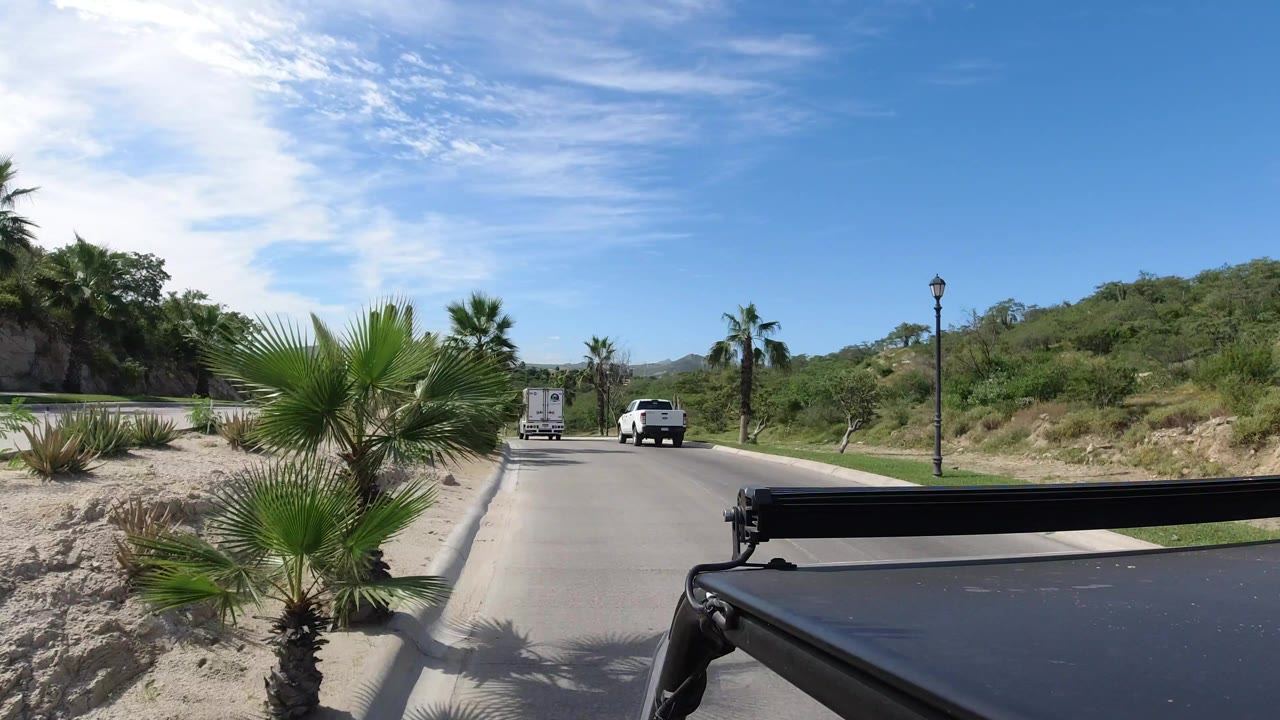 Forward-looking Los Cabos road view from a car, with a small palm in the foreground, two white vehicles ahead, green hills, blue sky, and no visible people.
