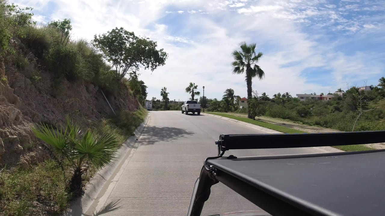 Vehicle-mounted Los Cabos road view with the lead truck farther along the lane, dry hillside plants on the left, palms on both sides, and no visible people.