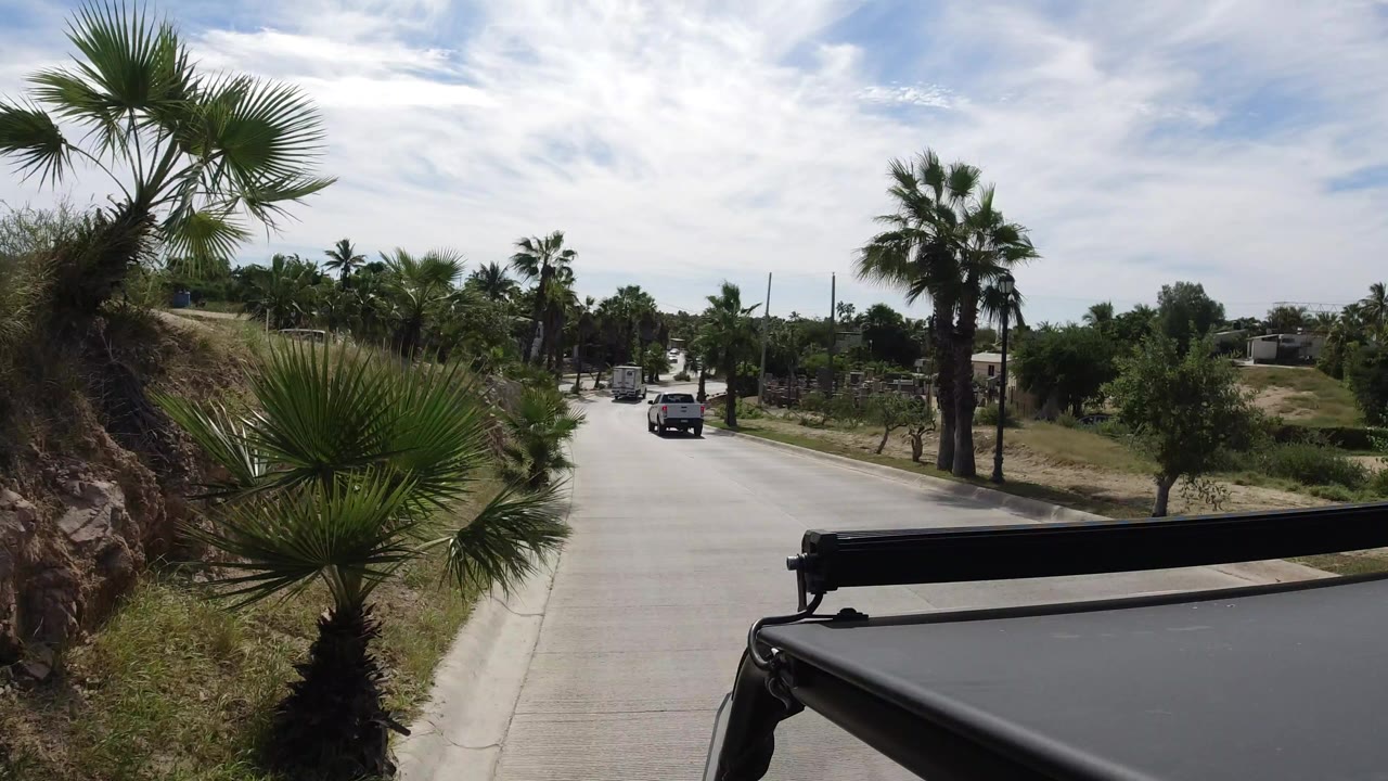 Opening frame in Los Cabos, Mexico, showing a pale road lined with palms and dry greenery, a dark vehicle roof edge in the foreground, a vehicle ahead, and no visible people.
