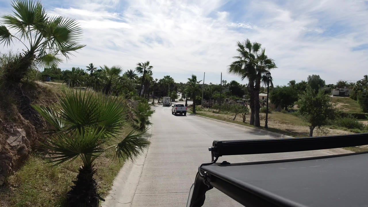 Early frame in Los Cabos, Mexico, with the road extending forward past palms, scrubby roadside planting, a distant vehicle, a dark foreground roof edge, and no visible people.