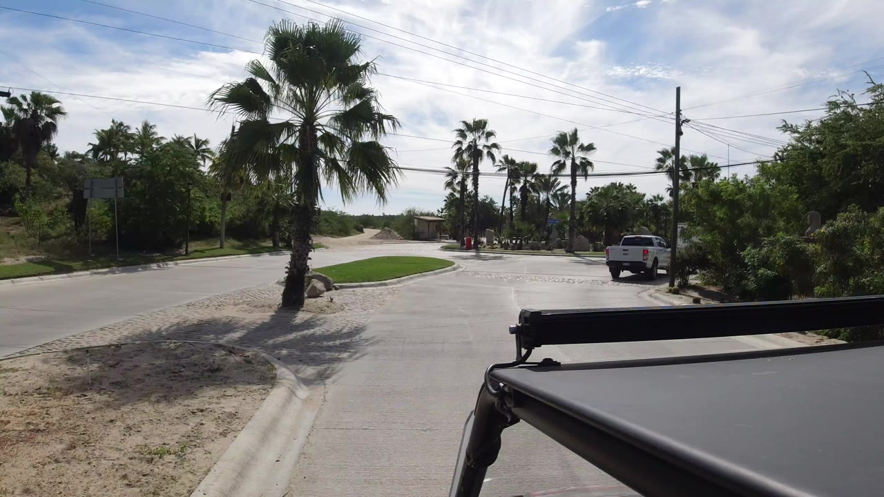 A vehicle-level view in Los Cabos shows a roundabout road with palms, grass, utility lines, and a white pickup ahead, with no visible people.