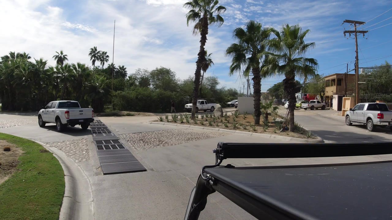 White pickup truck driving away on a Los Cabos road with palms, drainage grates, parked vehicles, low buildings, and no visible people.