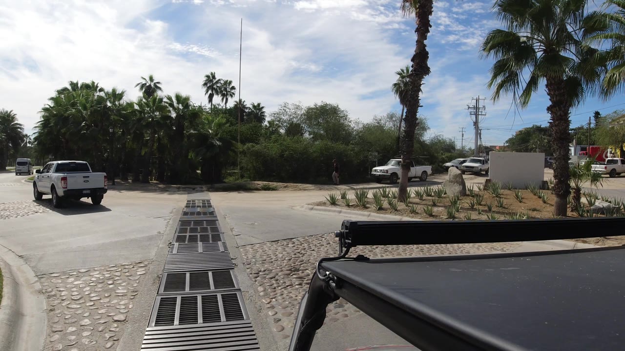 White pickup centered near drainage grates with palms and parked cars around the road.