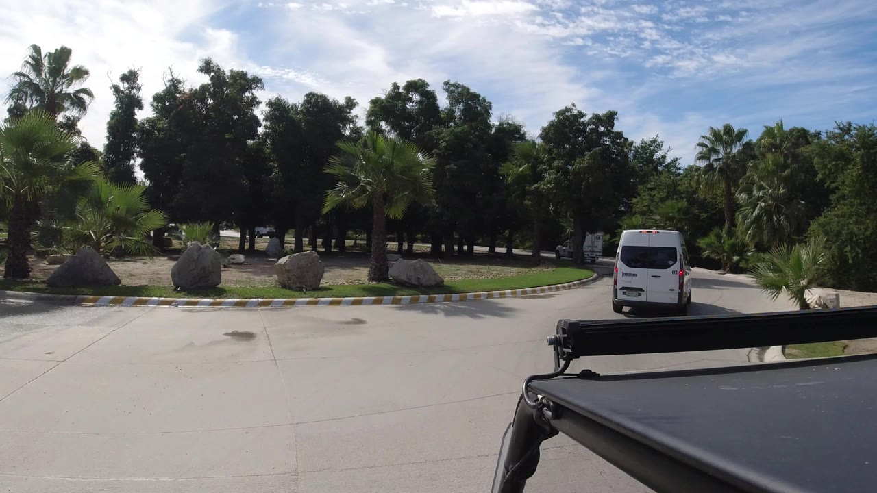 White van farther along the Los Cabos roadway, with a black foreground rail, landscaped grass, palms, shade trees, and bright sun across the pavement, with no visible people.
