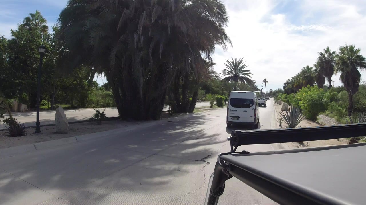 White van driving along a palm-lined road in Los Cabos, Mexico, with a dark vehicle or camera rig edge in the foreground and no visible people.