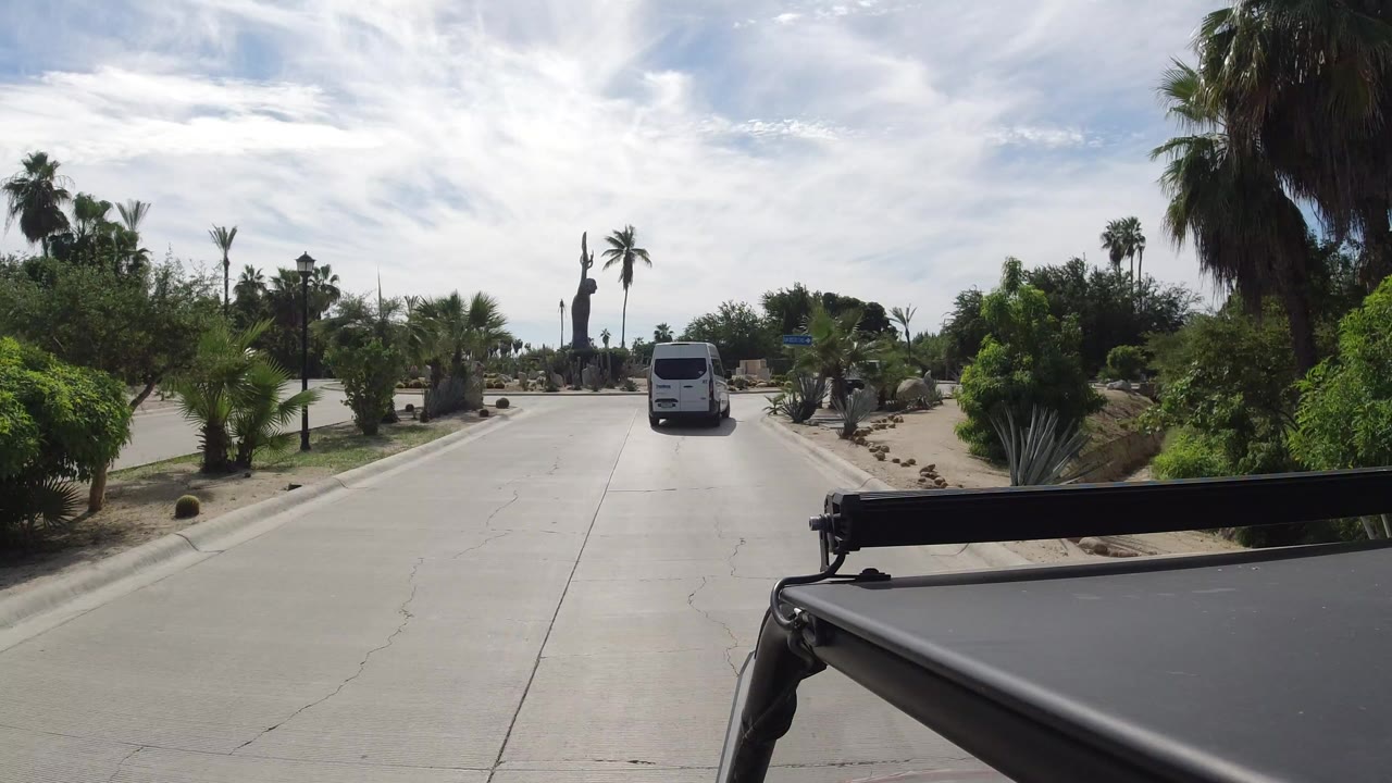 Vehicle-mounted view of a paved road with a white van ahead and landscaped palms.