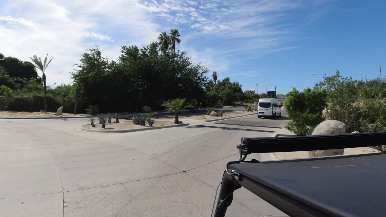 Opening view from a vehicle in Los Cabos, Mexico, showing a paved road, desert plants, palms, trees, and a white van ahead under bright blue sky, with no visible people.