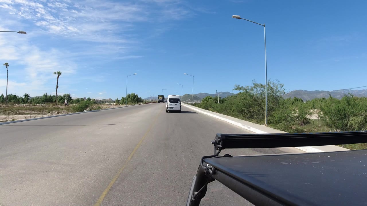 Los Cabos roadway seen from a vehicle-mounted angle, with a white van ahead, roadside greenery, lamp posts, distant mountains, blue sky, and no visible people.