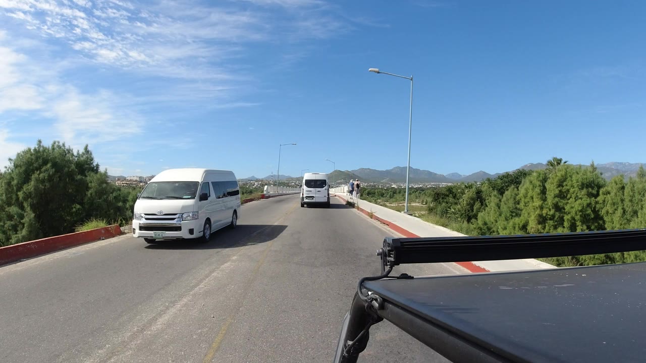 Vehicle-mounted view in Los Cabos, Mexico, with a white van centered ahead, a dark foreground vehicle edge, green bushes along the road, distant mountains, and no visible people.
