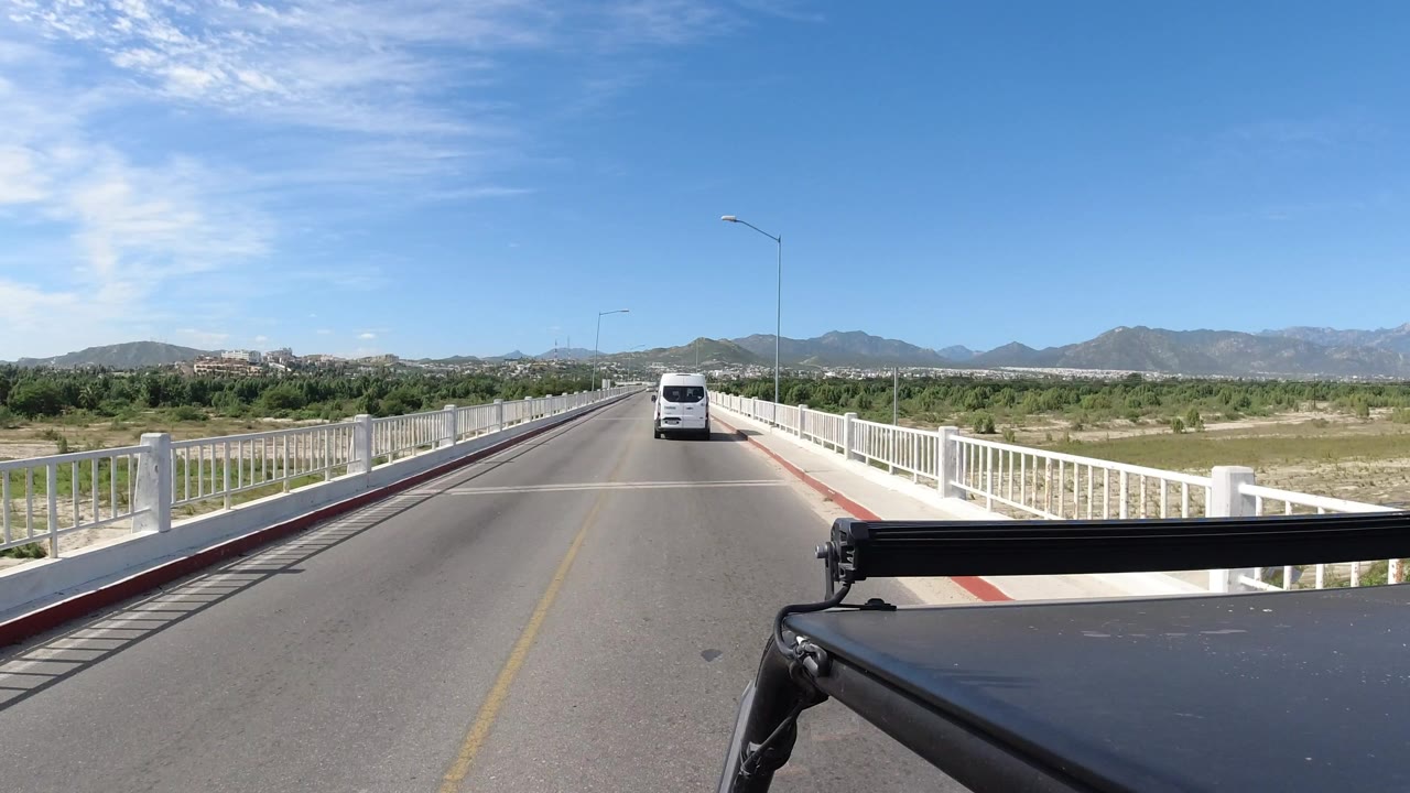 The Los Cabos bridge view continues with the white van centered farther up the two-lane road, white guardrails on both sides, scrubby green land, mountains, and no visible people.
