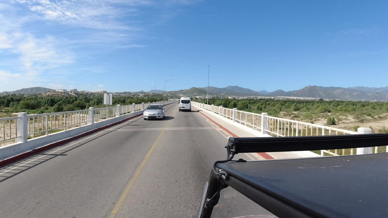 Opening frame of a Los Cabos bridge crossing from a vehicle viewpoint, with a white van ahead, an oncoming car, white guardrails, green vegetation, distant mountains, blue sky, and no visible people.