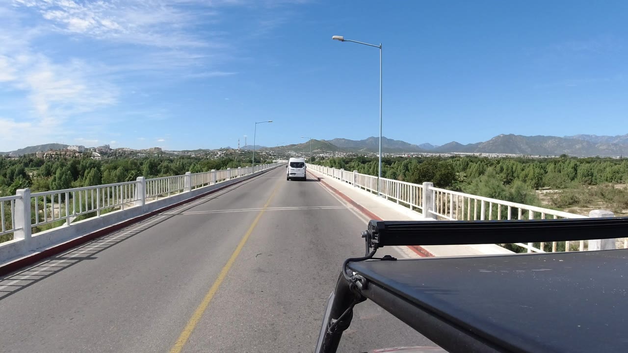 A centered road view in Los Cabos, Mexico, shows the van slightly farther along the elevated bridge, with white railings, roadside lights, trees, mountains, and no visible people.