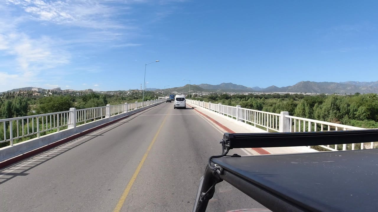 At the start of the Los Cabos clip, a forward-facing view shows a bridge road, white railings, a van ahead, green vegetation, distant mountains, bright blue sky, and no clearly visible people.