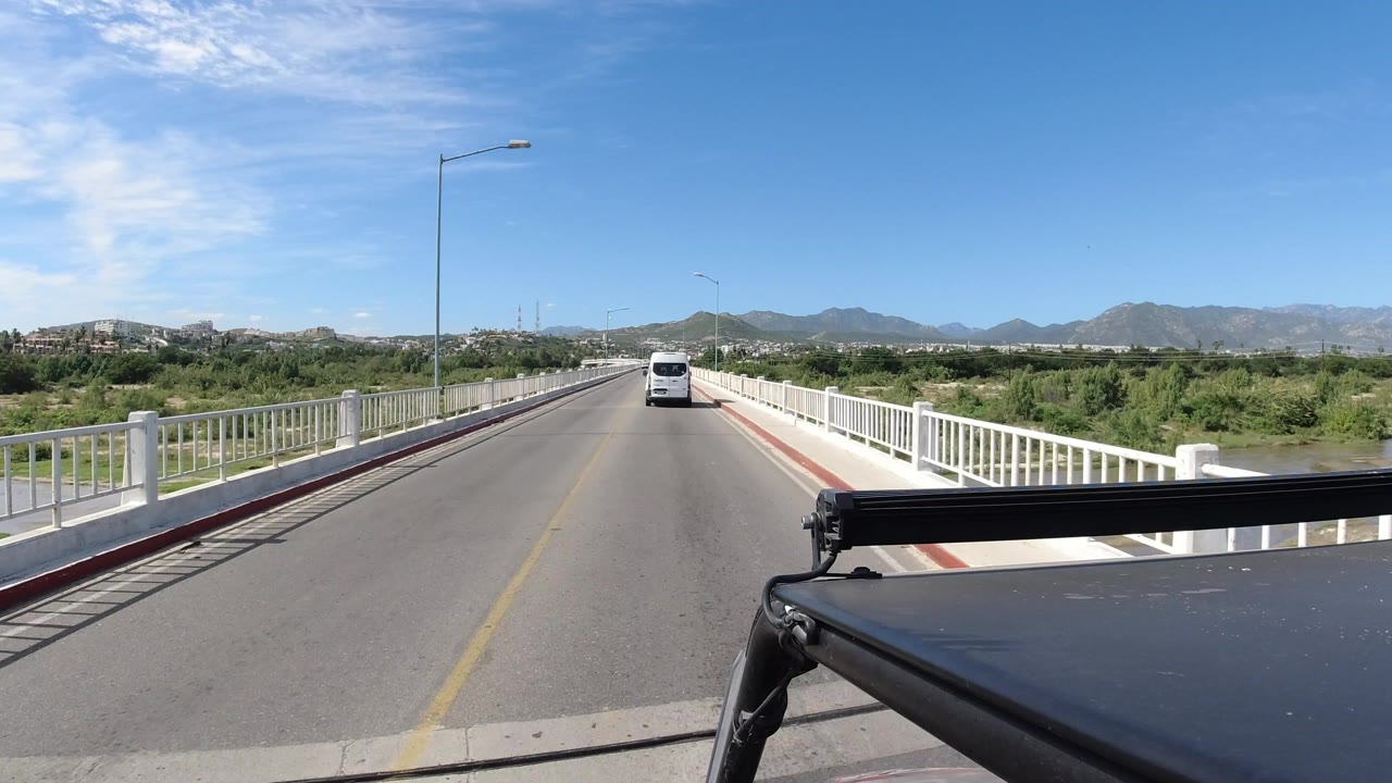 Los Cabos bridge crossing seen from a black vehicle, with the white van farther along the center lane, white railings on both sides, low mountains on the horizon, and no visible people.