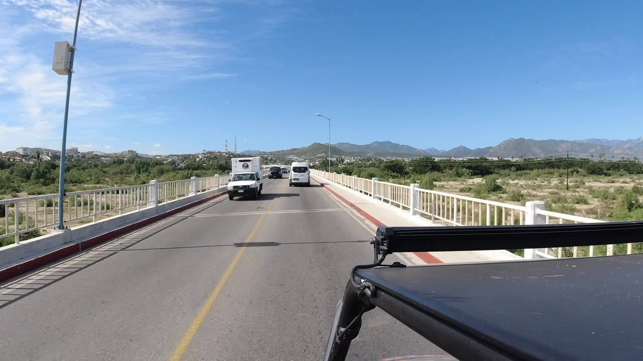 A Los Cabos bridge road with white railings, oncoming vans, scrub vegetation, distant mountains, bright sky, and no visible people.