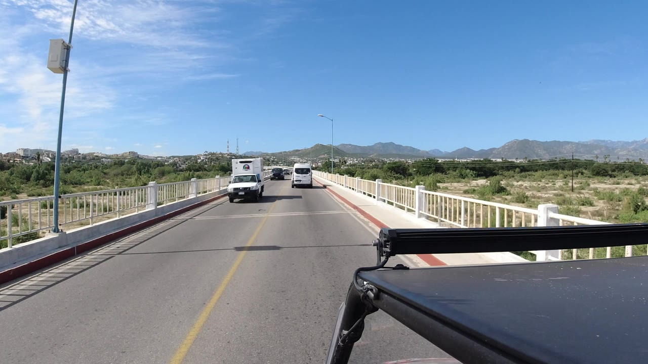 A forward road view in Los Cabos shows two oncoming vehicles, white bridge rails, scrubby land, mountains on the horizon, and no visible people.