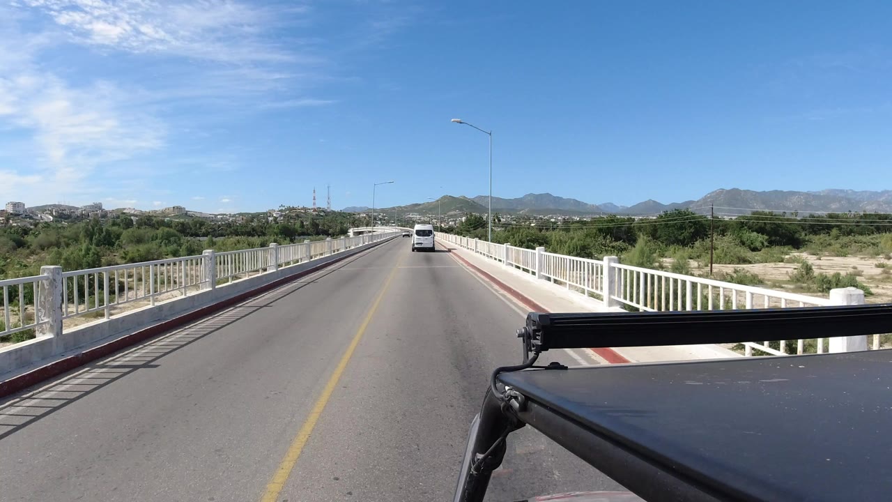 Opening view from a vehicle on a two-lane bridge in Los Cabos, Mexico, with white railings, a white car ahead, low mountains in the distance, and no visible people.