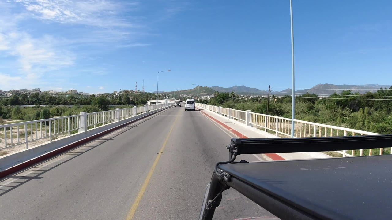 Two-lane bridge road with white railings, trees, distant hills, and a car ahead.