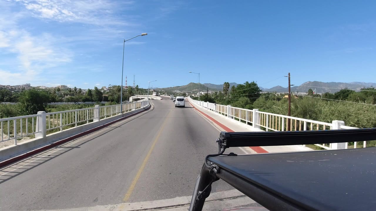 Roof-mounted car POV on a bridge road with white railings and hills ahead.